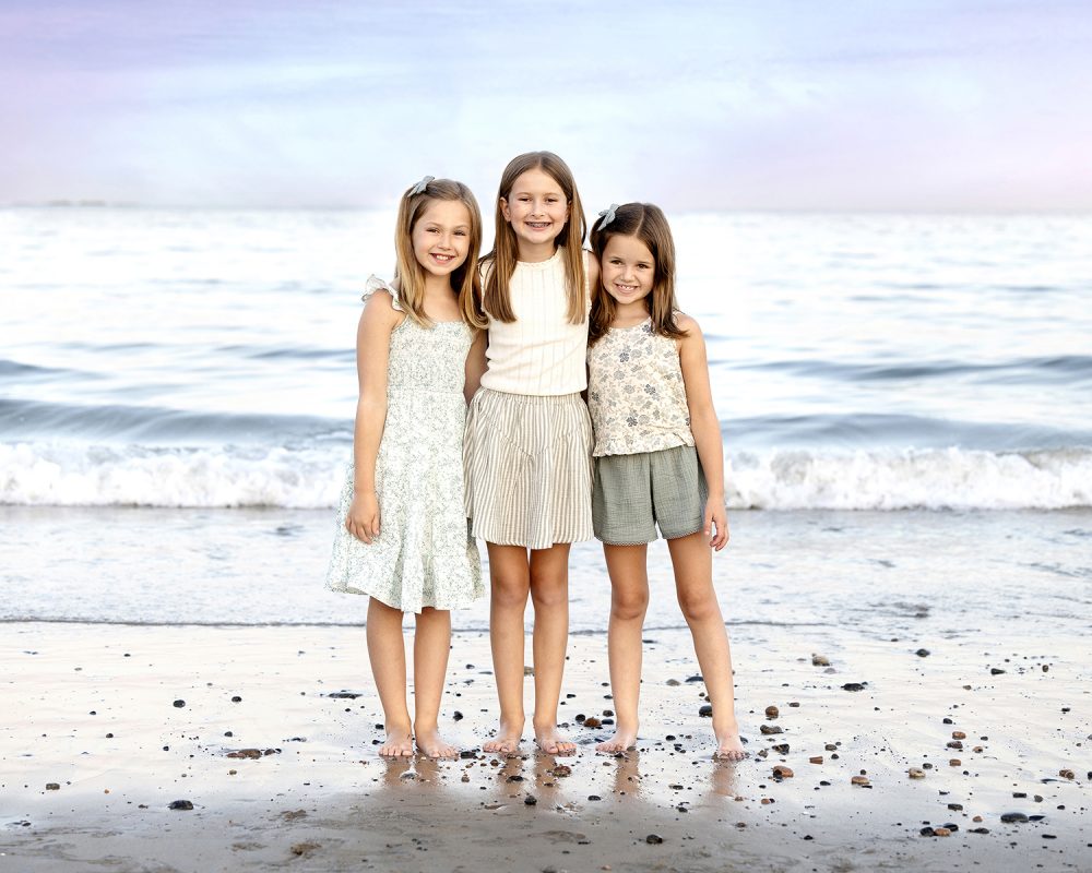 Three sisters standing together at the shoreline during a summer beach portrait session photographed by a Massachusetts family photographer